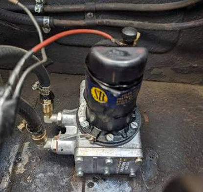 A black domed end cover carrying a yellow and blue SU badge, mounted on an alloy body with several pipe unions, a red electrical feed and braided hoses, against a dark painted panel