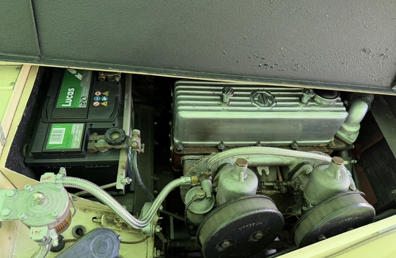 Engine bay of a cream MG TF 1500 seen from the carburettor side, showing the twin SUs with pancake air filters, the alloy rocker cover, the Lucas battery and the braided fuel line