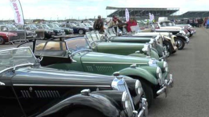 A line-up of MG T-Type roadsters — black, green and cream cars parked nose-out on a paddock at a British classic car meeting, with owners and enthusiasts standing between the cars and grandstands in the background.