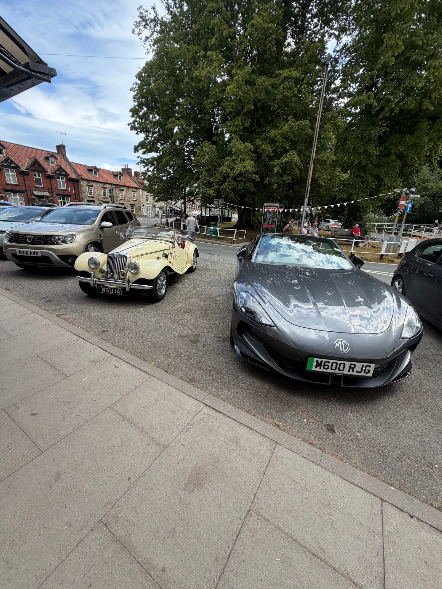 The MG TF1500 WSU 141 parked in the centre of Thornton-le-Dale beside a modern grey MG Cyberster electric roadster, with the village's great chestnut tree and stone cottages behind.
