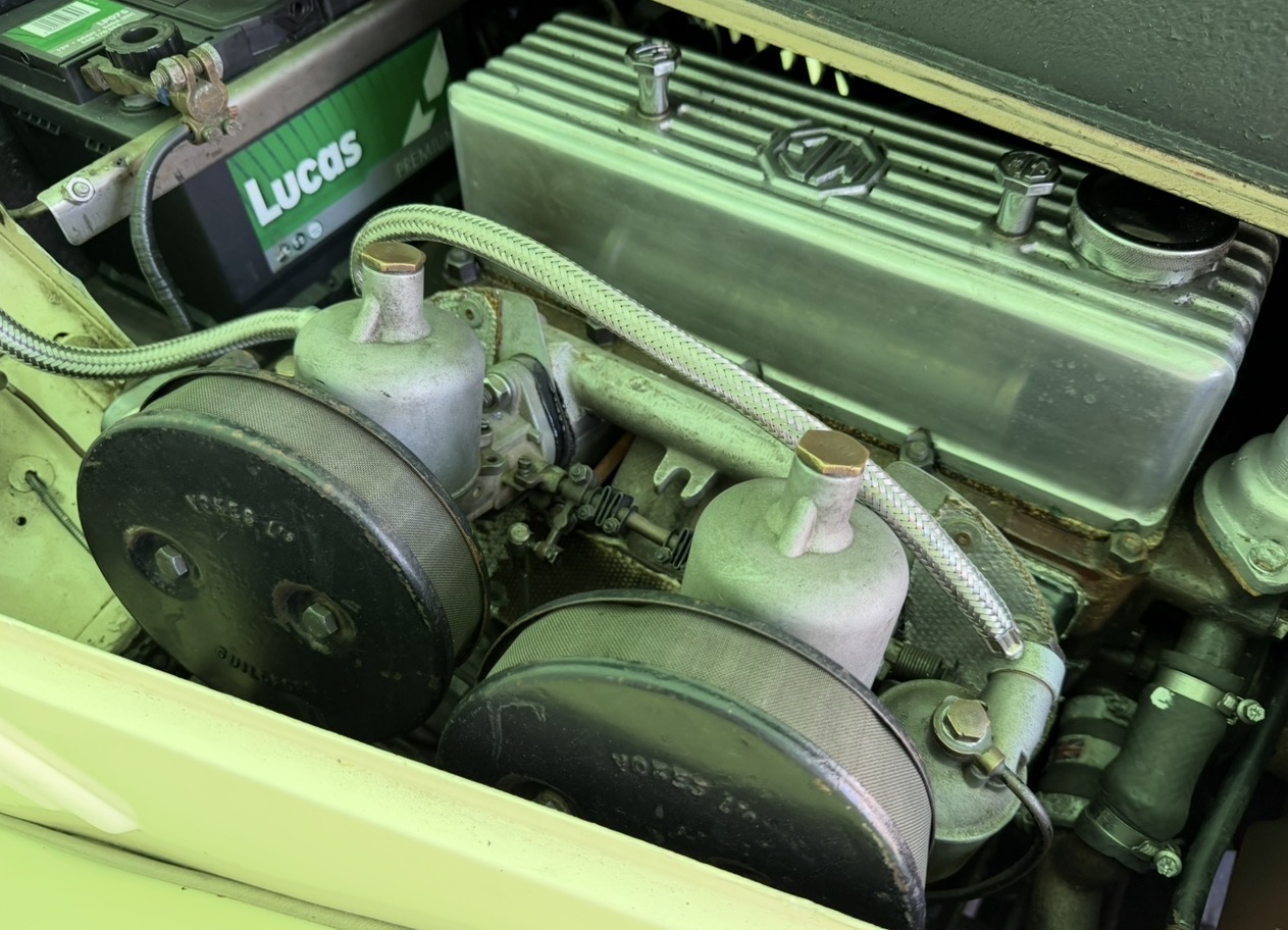 Close view of the twin SU carburettors on a cream MG TF 1500 with Vokes pancake air filters fitted, the throttle linkage and return spring between them, and braided fuel pipes to each float chamber
